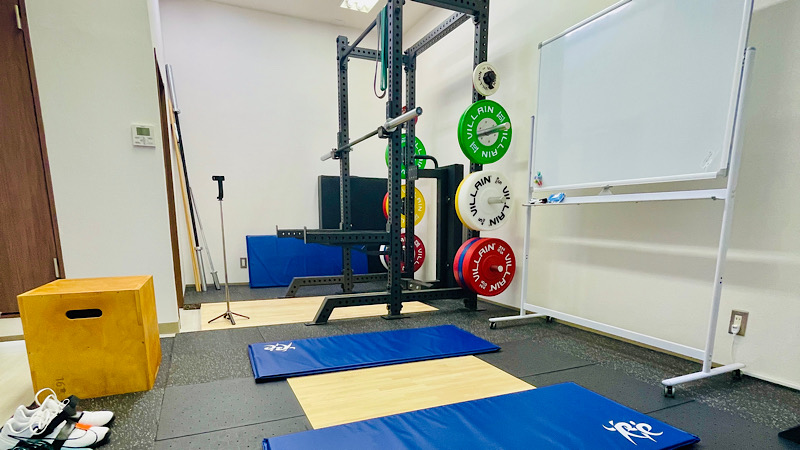 トレーニングスペース。中央には2枚のストレッチマットが敷いてあり、壁際にはホワイトボードが置かれて、奥にはパワーラックが設置されている。
