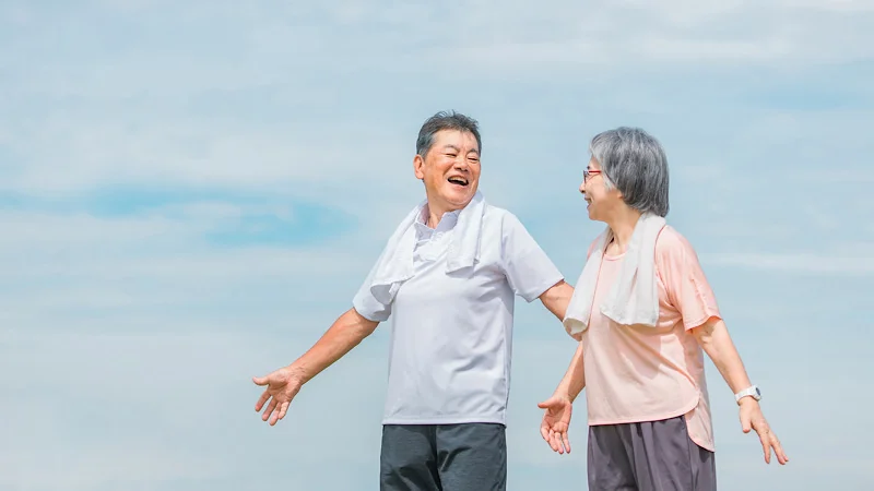 運動後、首に白いタオルを巻いて晴れやかな表情で笑いあう、アクティブな装いの高齢男女。背景には澄み渡った青空が広がっている