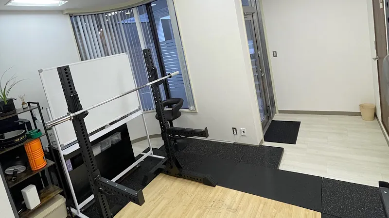 明るい日差しが差し込む清潔感のあるTAKASE GYMの内観。10kgバーベルが設置されたスクワットラックの後ろには説明に使うホワイトボードが置かれている。