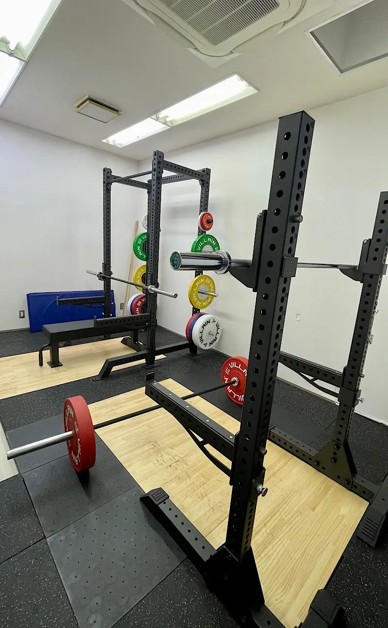 木目調のプラットフォームが設置された清潔感のあるジム内装。手前にはスクワットラック、奥にはベンチプレス用のハーフラックが並び、床にはバーベルが準備されています。