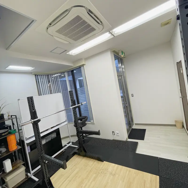 明るい日差しが差し込む清潔感のあるTAKASE GYMの内観。10kgバーベルが設置されたスクワットラックの後ろには説明に使うホワイトボードが置かれている。